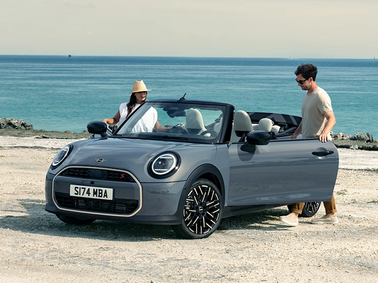 Frontansicht eines MINI Cooper Cabrio in Copper Grey an einem Strand mit zwei daneben stehenden Personen.