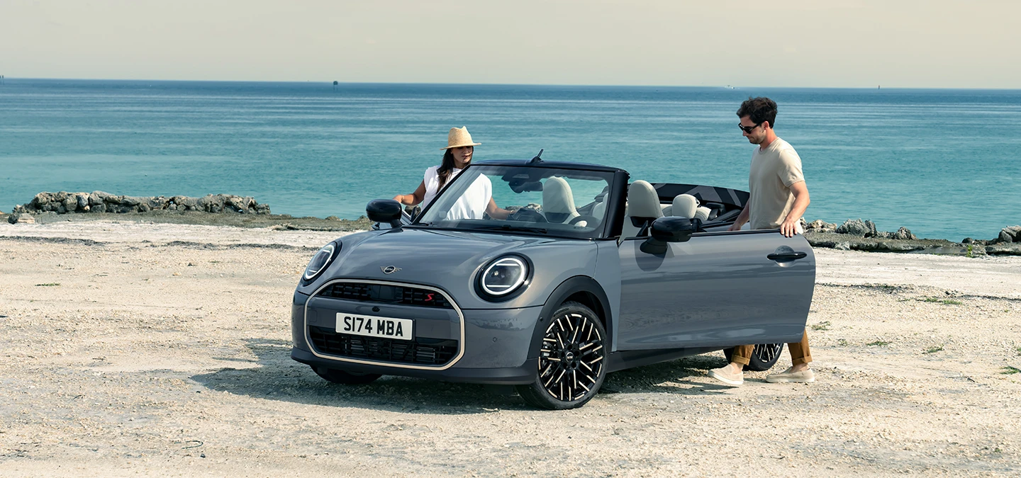 Frontansicht eines MINI Cooper Cabrio in Copper Grey an einem Strand mit zwei daneben stehenden Personen.