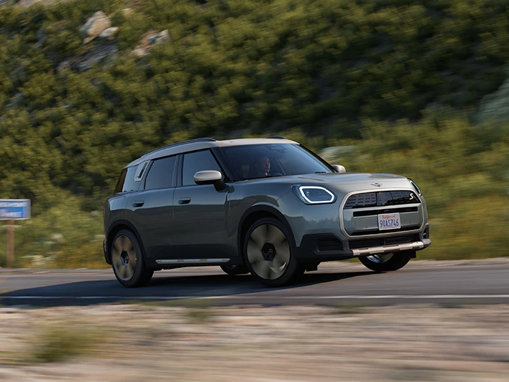 Seitenansicht eines auf einer holprigen Strasse fahrenden vollelektrischen MINI Countryman in Smokey Green mit Dach in Vibrant Silver.