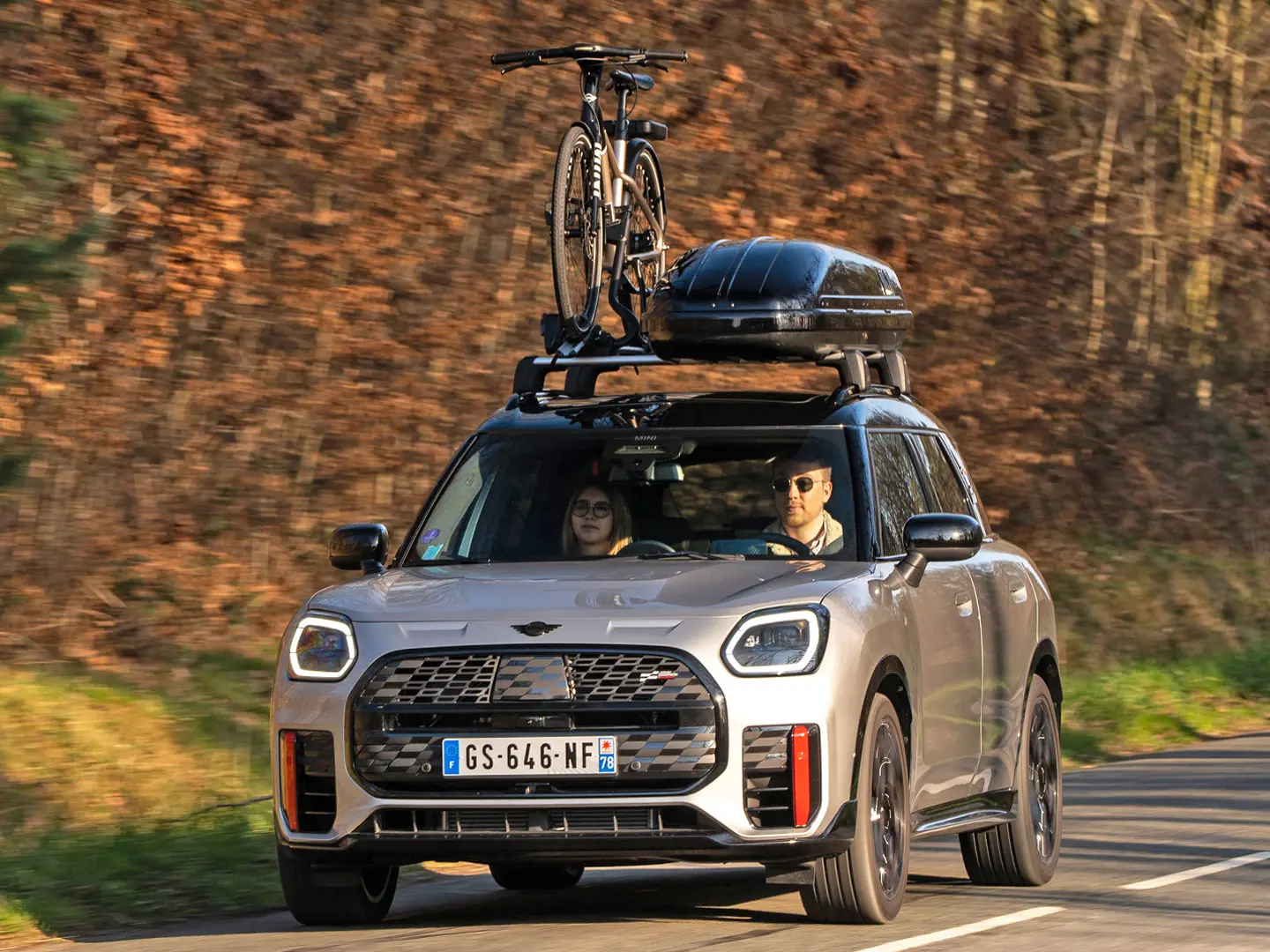 MINI Countryman Cooper in Silver su una strada di montagna con portapacchi MINI, box per tetto MINI, portabici da tetto MINI con una bici montata e aletta MINI John Cooper Works – vista anteriore. 