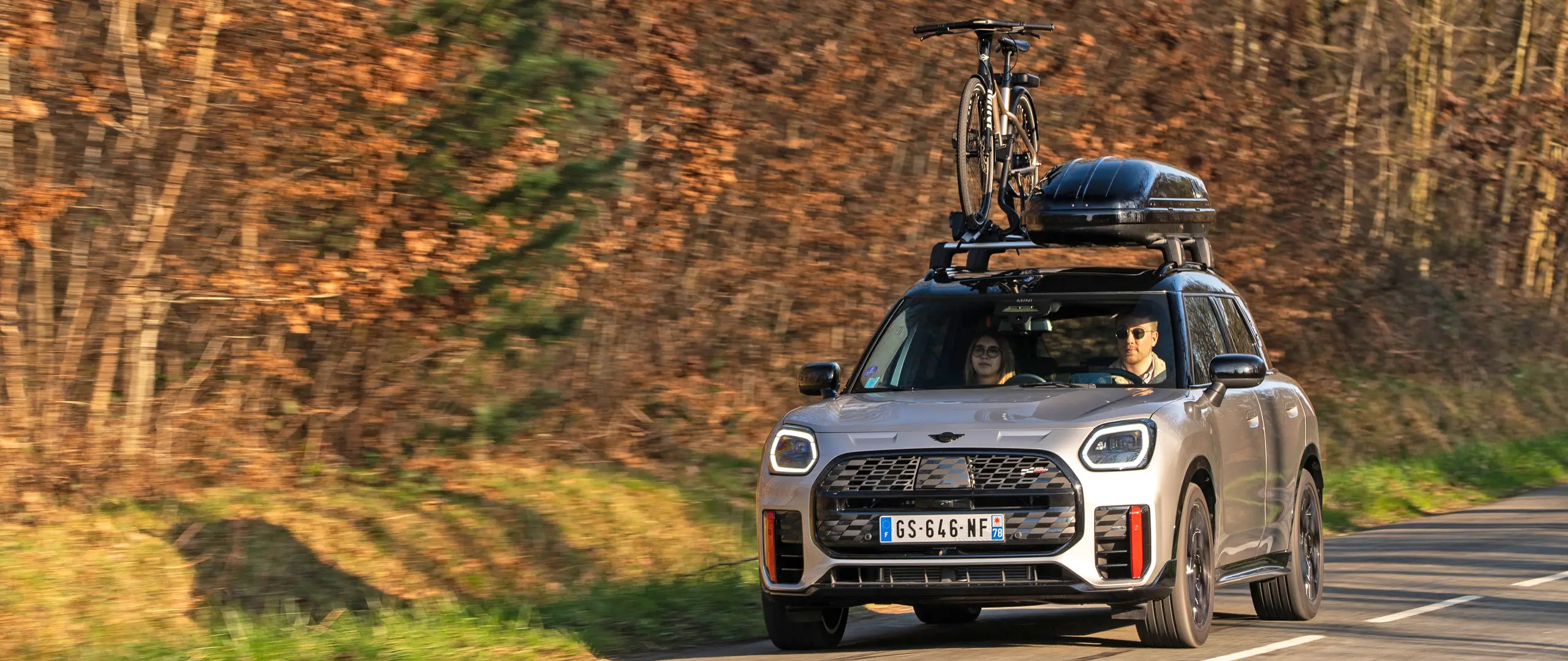 MINI Countryman Cooper in Silver su una strada di montagna con portapacchi MINI, box per tetto MINI, portabici da tetto MINI con una bici montata e aletta MINI John Cooper Works – vista anteriore. 