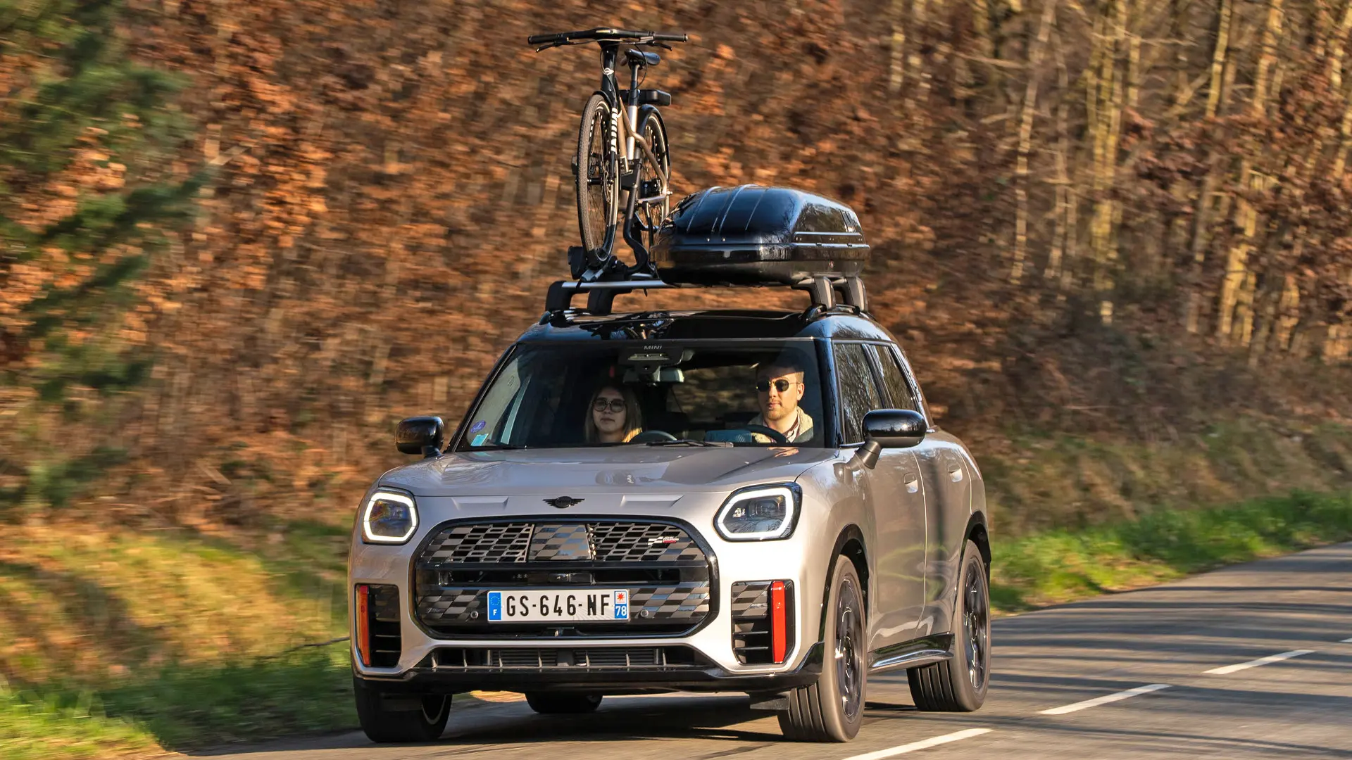 MINI Countryman Cooper in Silver su una strada di montagna con portapacchi MINI, box per tetto MINI, portabici da tetto MINI con una bici montata e aletta MINI John Cooper Works – vista anteriore. 