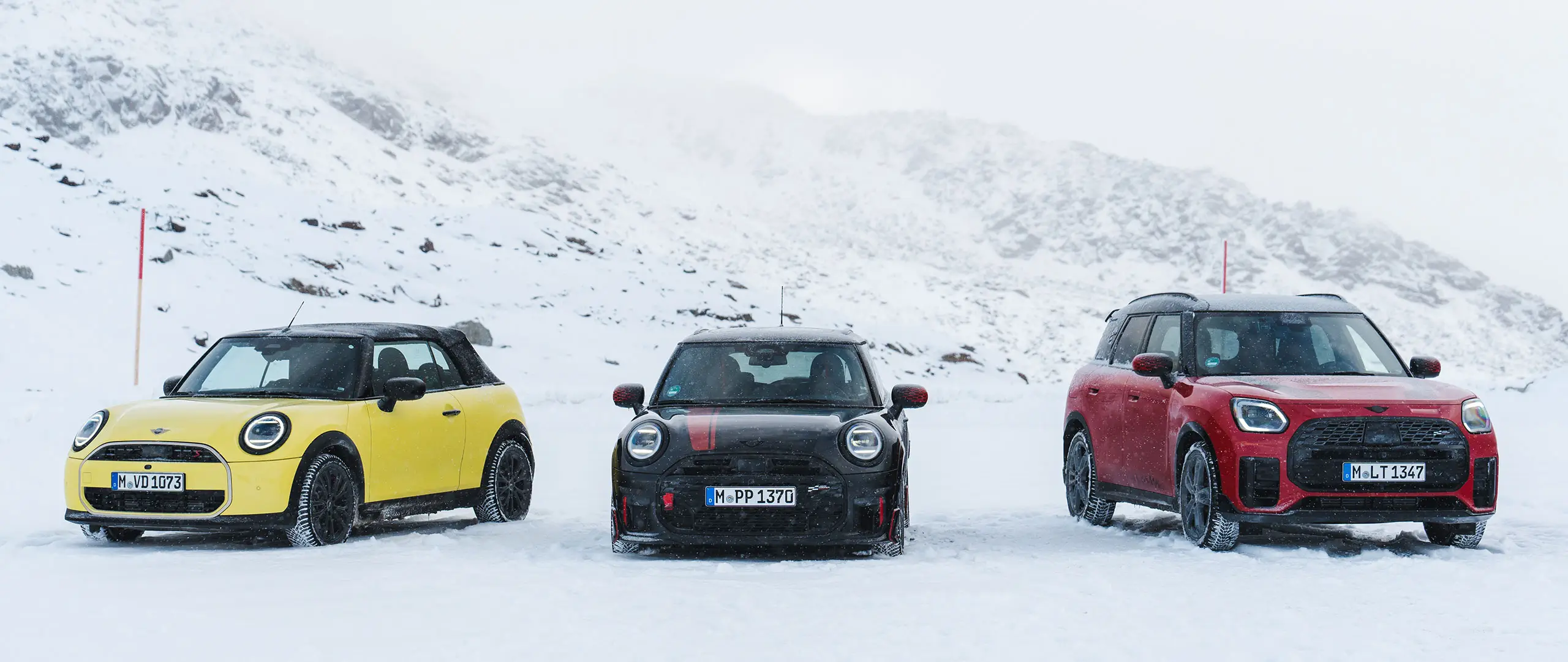 MINI Cooper Cabrio in Sunny Side Yellow, MINI John Cooper Works in Midnight Black II e MINI Countryman Cooper in Chilli Red – ferme su una strada di montagna innevata, vista anteriore.