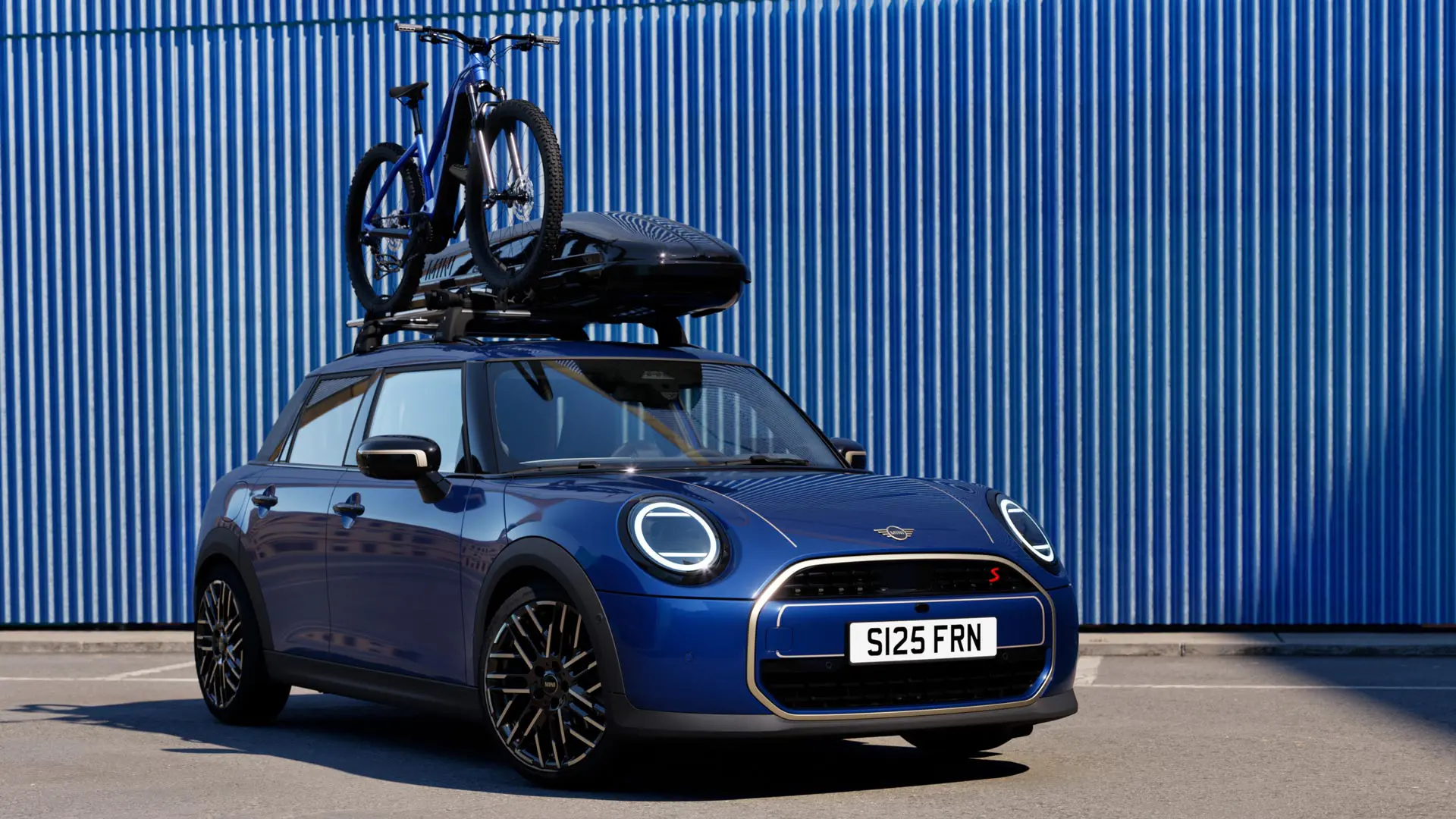 Vue de face de la MINI Aceman en Indigo Sunset Blue avec porte-vélos de toit MINI, transportant un vélo de montagne et un coffre de toit MINI 320.