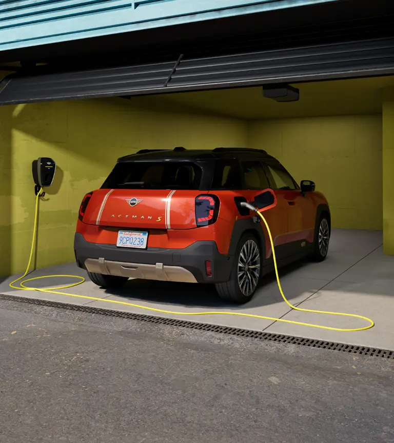 Heckansicht eines vollelektrischen MINI Aceman, der in einer Garage parkiert ist und mit der MINI Wallbox Plus geladen wird.