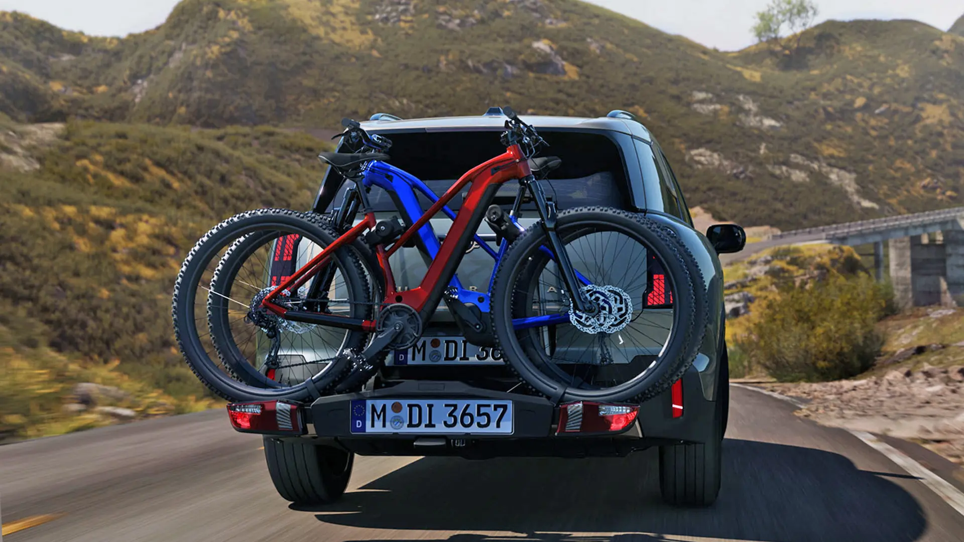 Heckansicht eines MINI Countryman in Smokey Green mit Veloheckträger und zwei Mountainbikes, der auf einer Bergstrasse fährt.
