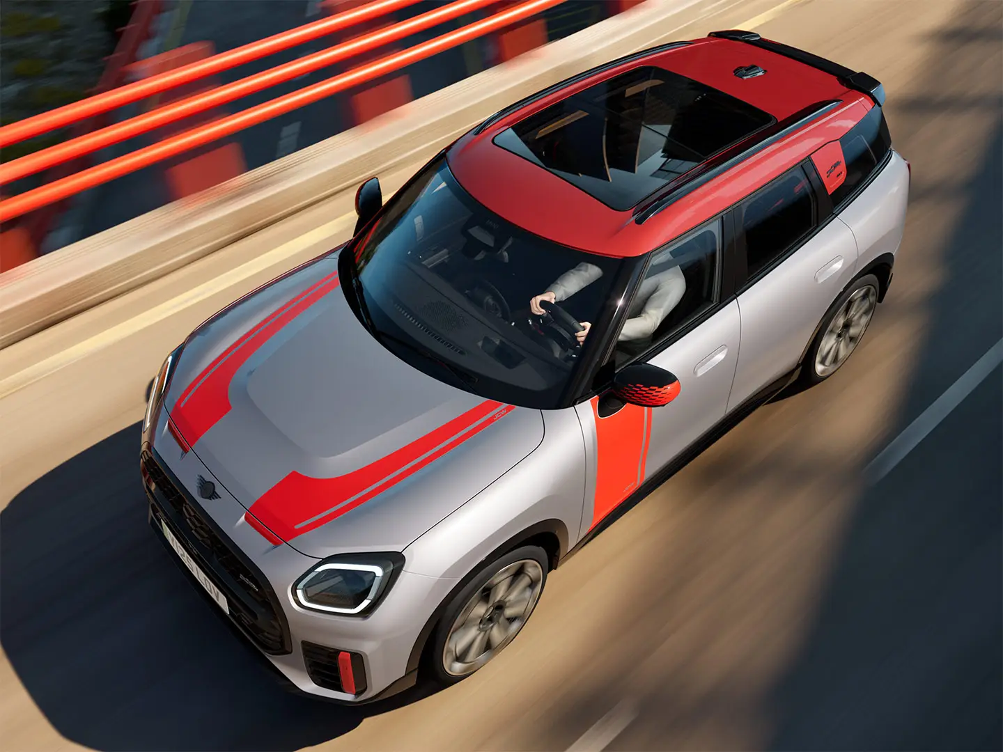 MINI Countryman John Cooper Works in Melting Silver con calotte degli specchietti retrovisori, decalcomanie laterali e sul cofano, tetto e alette in Chili Red, in marcia lungo una strada cittadina – vista frontale laterale.
