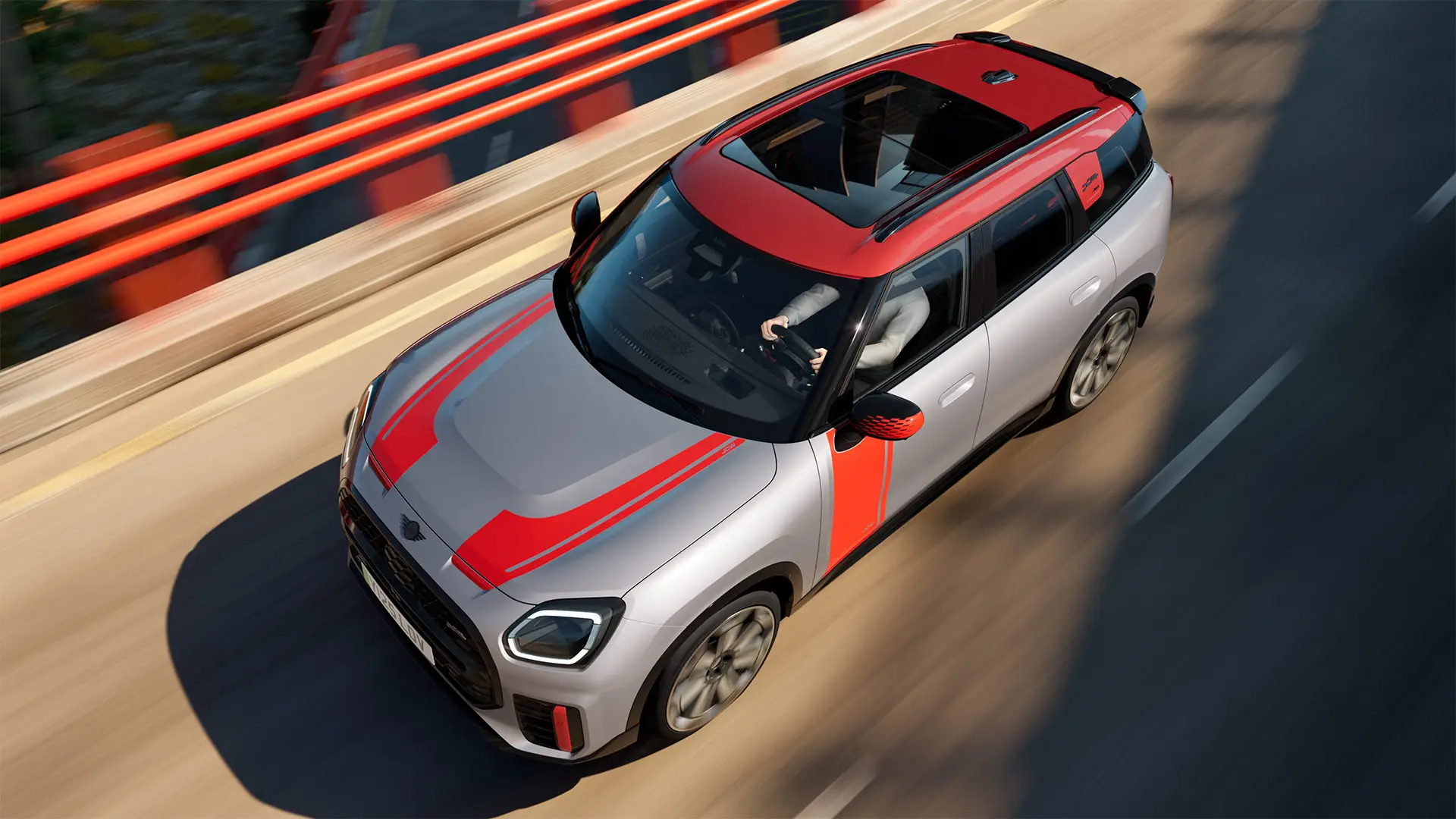 MINI Countryman John Cooper Works in Melting Silver con calotte degli specchietti retrovisori, decalcomanie laterali e sul cofano, tetto e alette in Chili Red, in marcia lungo una strada cittadina – vista frontale laterale.