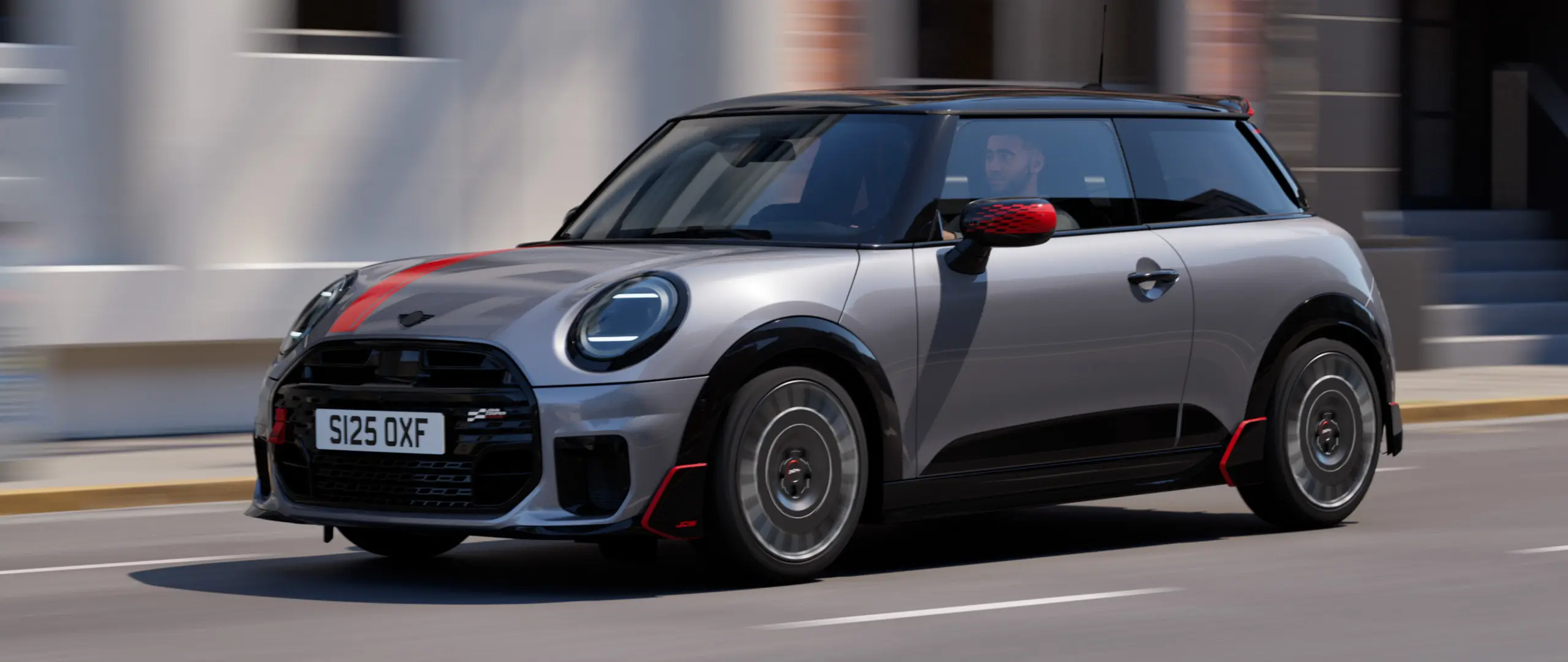 MINI Cooper a 3 porte in Melting Silver con calotte degli specchietti retrovisori John Cooper Works e decalcomania sul cofano in Chili Red, spoiler sul tetto e alettone in nero, mentre percorre una strada cittadina – vista frontale laterale.