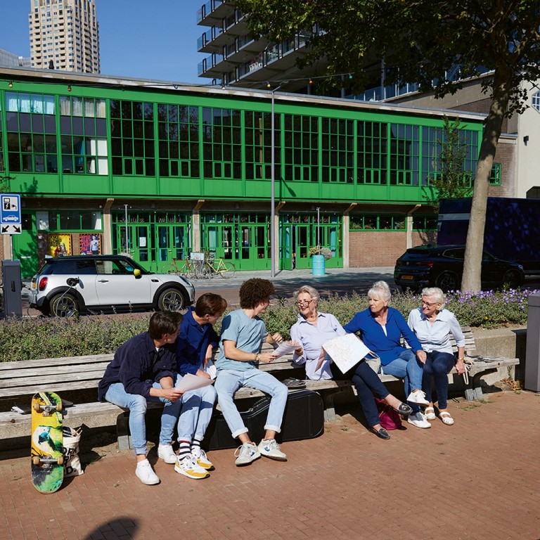 Trois jeunes discutent avec trois dames âgées sur le banc d’un parc, derrière se gare une MINI Electric