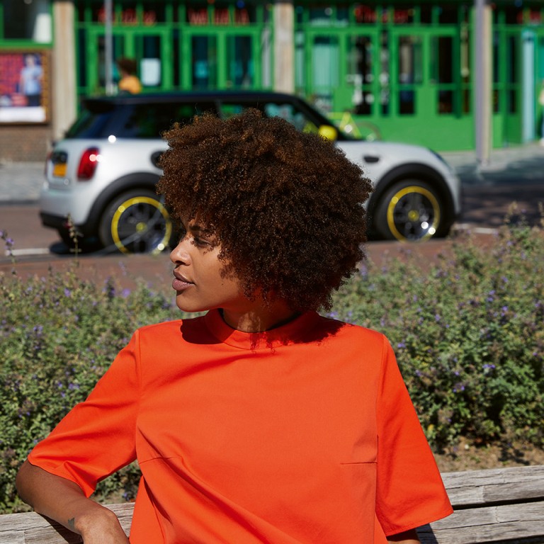 Une femme est assise sur un banc et regarde sur le côté, derrière elle une MINI Electric se gare