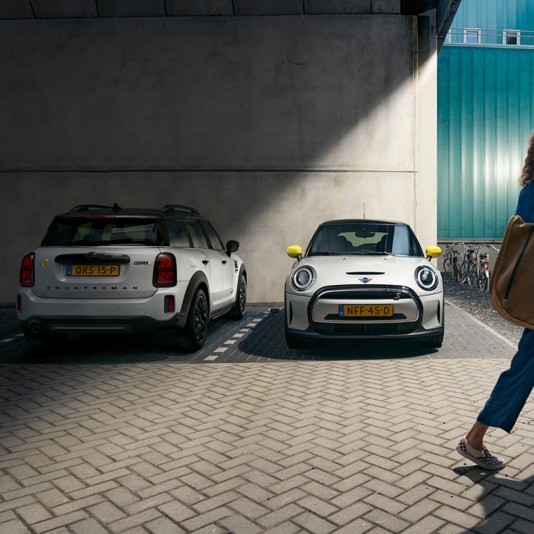 MINI Electric und MINI Plug-in-Hybrid Frau läuft an einem geparkten MINI Electric und einem geparkten MINI MINI Plug-in-Hybrid vorbei