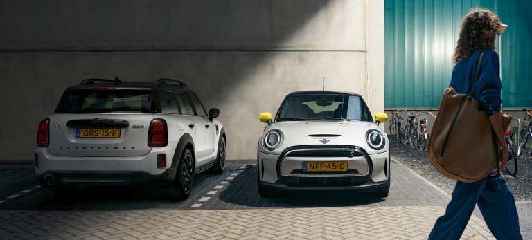 MINI Electric et MINI plug-in-hybrid Une femme passe à côté d’une MINI Electric garée et d’une MINI plug-in-hybrid garée